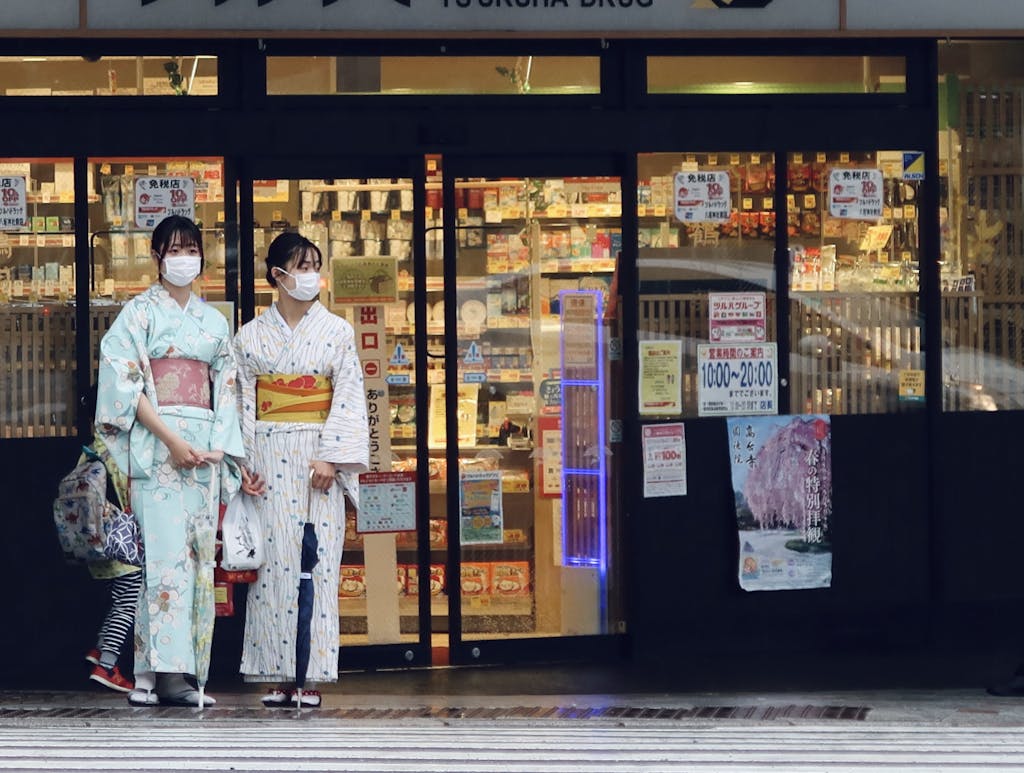 Two Asian women in kimonos wearing masks in front of a Japanese market.
